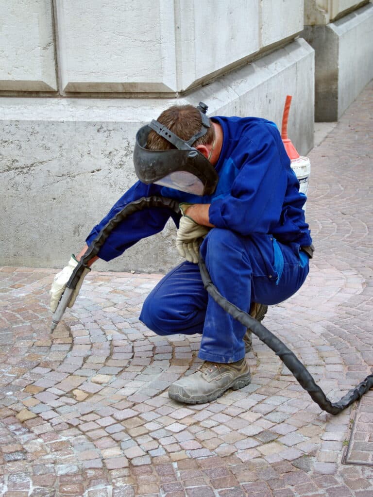 Nettoyage de pavés : Ouvrier spécialisé en action Ouvrier en combinaison bleue, masque de protection, nettoie les pavés d'une rue à l'aide d'un outil de sablage.
