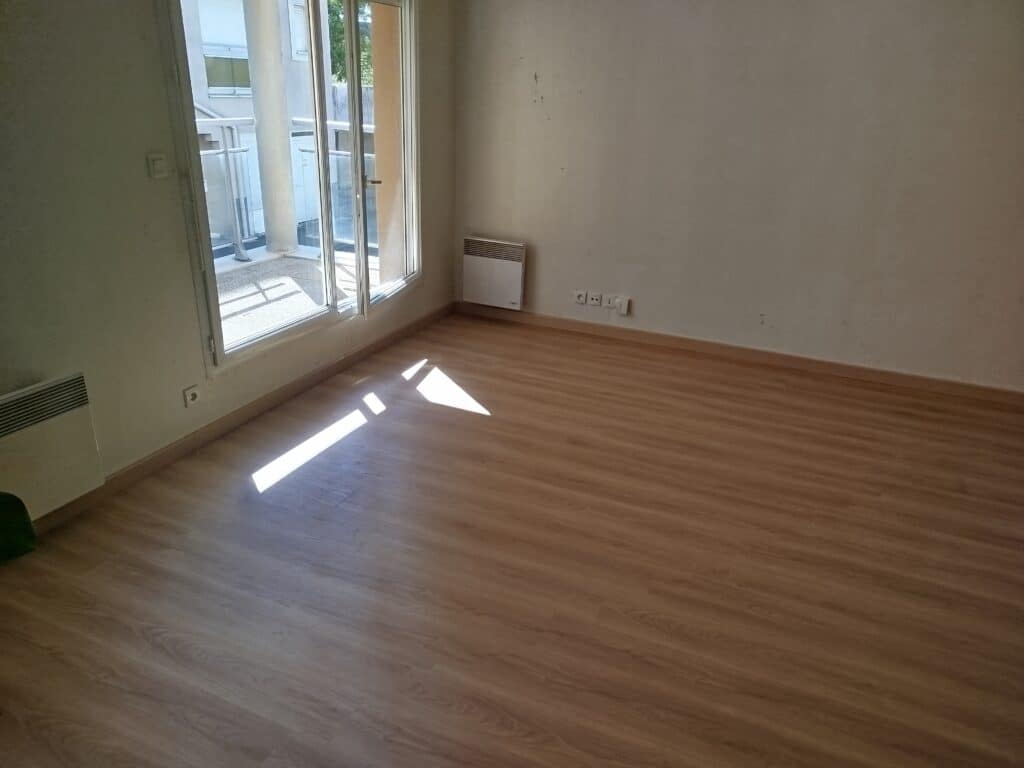 Pièce vide lumineuse d'appartement avec parquet clair, grande fenêtre donnant sur un balcon et radiateurs muraux. Belle lumière naturelle.