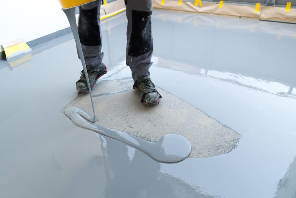 Ouvrier verse une résine grise sur un sol en béton, portant des chaussures à pointes pour l'application d'un revêtement époxy.