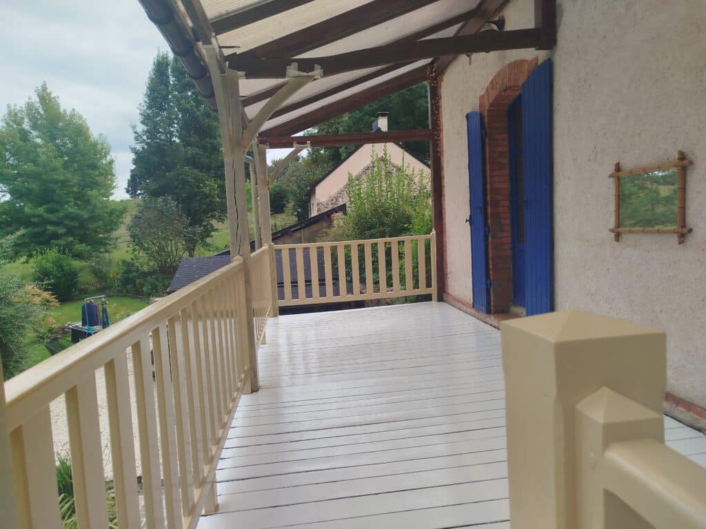 Grand balcon couvert en bois clair et blanc, avec vue sur un jardin verdoyant, une maison et des arbres. Porte bleue de la propriété.