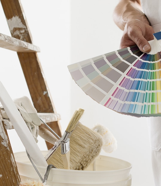 Main choisissant des couleurs avec un nuancier de peinture. Au premier plan, pinceaux, rouleau, seau et échelle de rénovation.