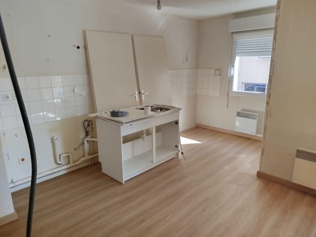 Cuisine vide en rénovation avec meuble d'évier, carrelage mural blanc, parquet flottant clair, fenêtre et radiateur.