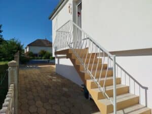 Escalier extérieur jaune clair avec rambarde en fer forgé blanc vers une maison blanche. Jardin et pavés hexagonaux sous soleil.
