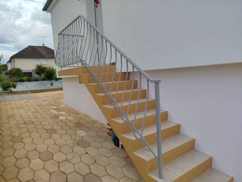 Escalier extérieur en béton ocre et carrelage clair avec rampe métallique grise. Devant une maison blanche et des pavés hexagonaux.