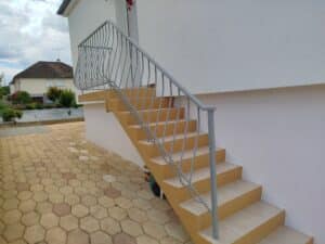 Escalier extérieur en béton ocre et carrelage clair avec rampe métallique grise. Devant une maison blanche et des pavés hexagonaux.