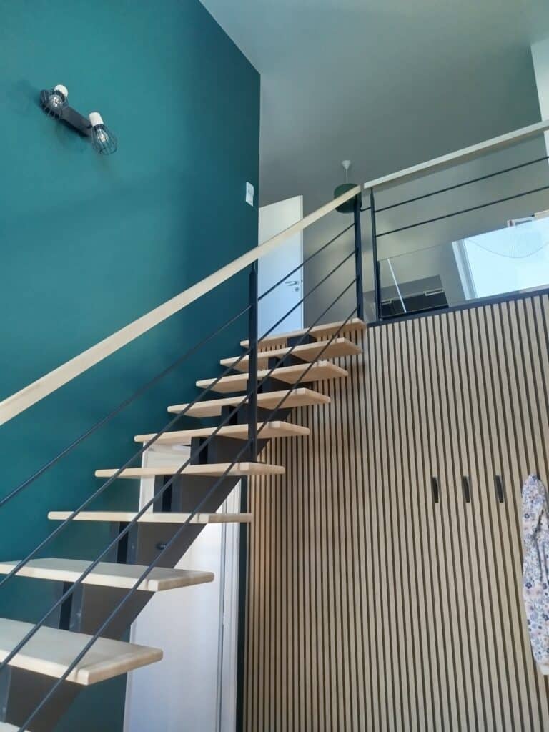 Escalier flottant bois et métal sur mur bleu canard, avec luminaire industriel. Parement mural latté bois clair et placards intégrés.