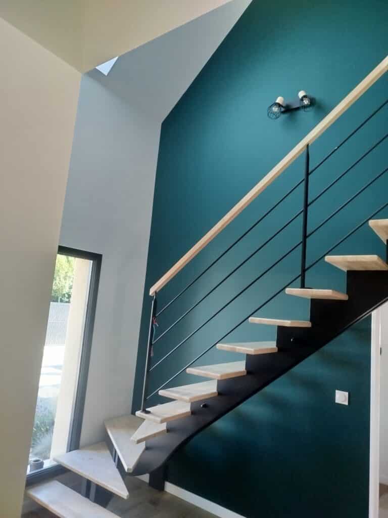 Escalier moderne en bois clair et métal noir sur mur bleu canard. Rampe bois, balustrade métal. Grande fenêtre lumière naturelle.