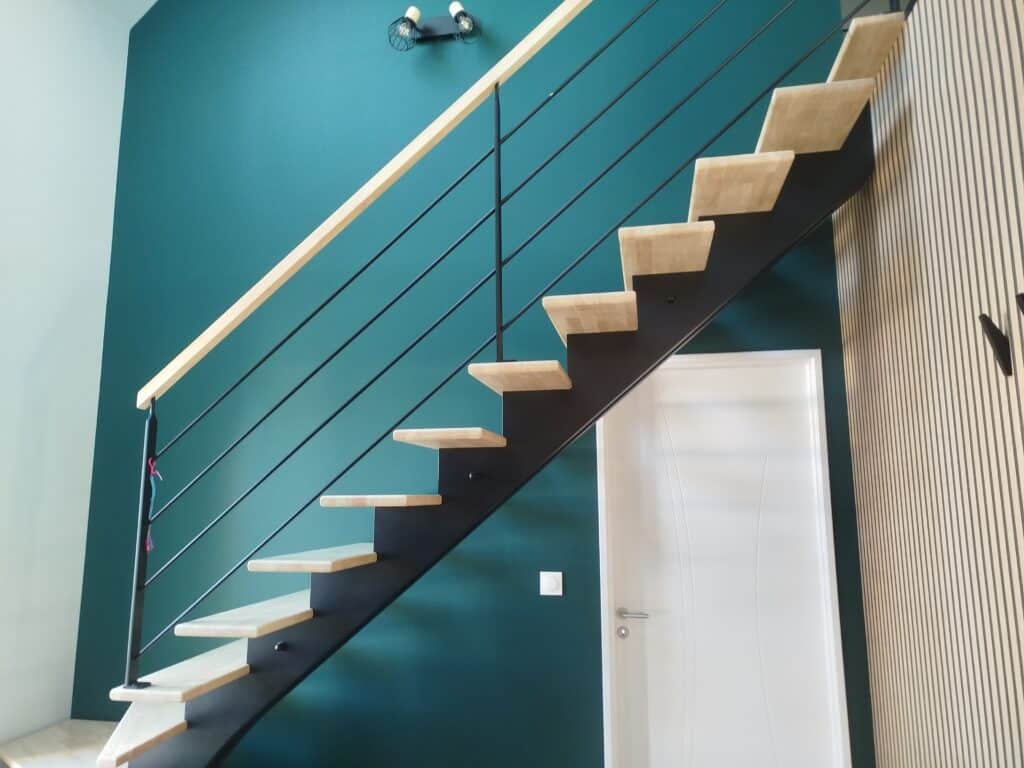 Escalier moderne avec marches en bois, rampe métallique noire, mur vert canard et parement mural à lattes.