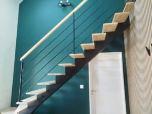 Escalier moderne avec marches en bois, rampe métallique noire, mur vert canard et parement mural à lattes.
