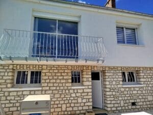 Façade de maison deux étages: pierre beige, enduit blanc, balcon avec garde-corps. Porte N°5 ouverte, fenêtres. Boîte aux lettres "PROTÉGÉ PAR". Reflet d'une personne.