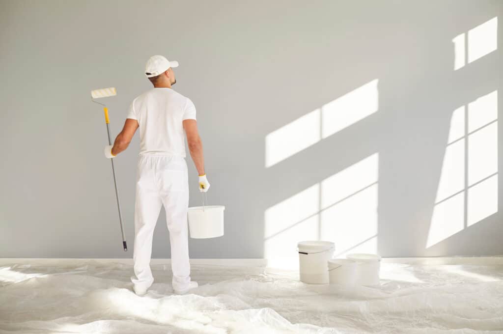 Peintre en blanc, dos tourné, tenant rouleau et seau, prêt à repeindre un mur gris. Ombres de fenêtre sur le mur et sol protégé.