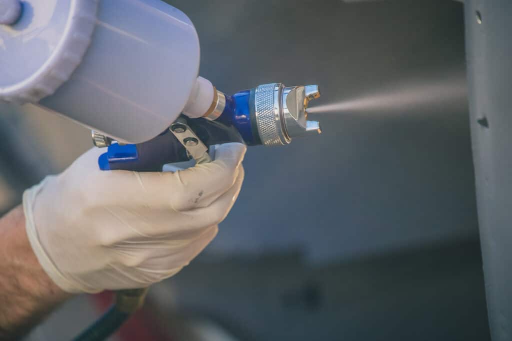 Pistolet à Peinture Professionnel: Application Précise Main gantée utilise un pistolet pulvérisateur bleu et blanc, projetant une brume fine. Application de peinture ou vernis.