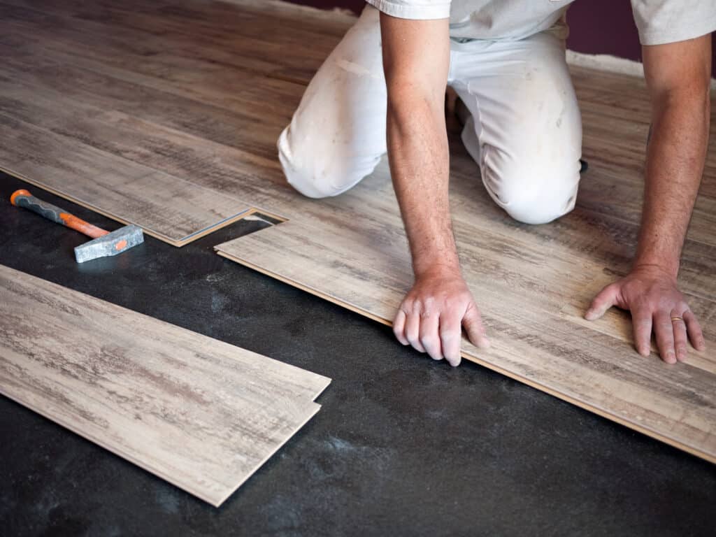 Homme posant du parquet stratifié clair sur un sous-plancher noir. Ses mains guident une lame tandis qu'un marteau repose à côté.