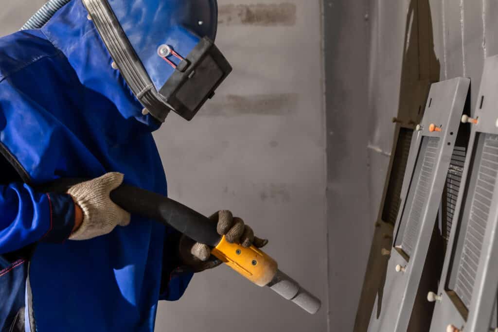 Ouvrier en combinaison bleue et casque de protection effectuant un sablage sur des pièces métalliques avec un pistolet industriel.