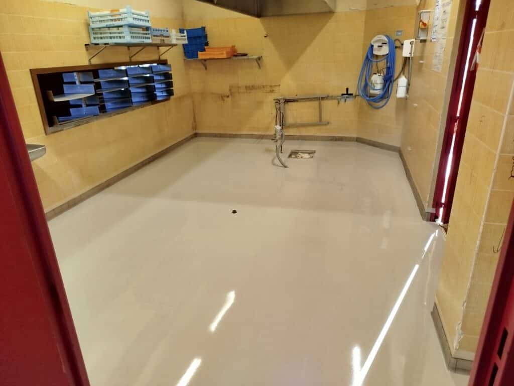 Salle professionnelle propre, sol en résine gris clair brillant et murs carrelés jaunes. Étagères avec caisses, équipement de nettoyage mural et drain central.