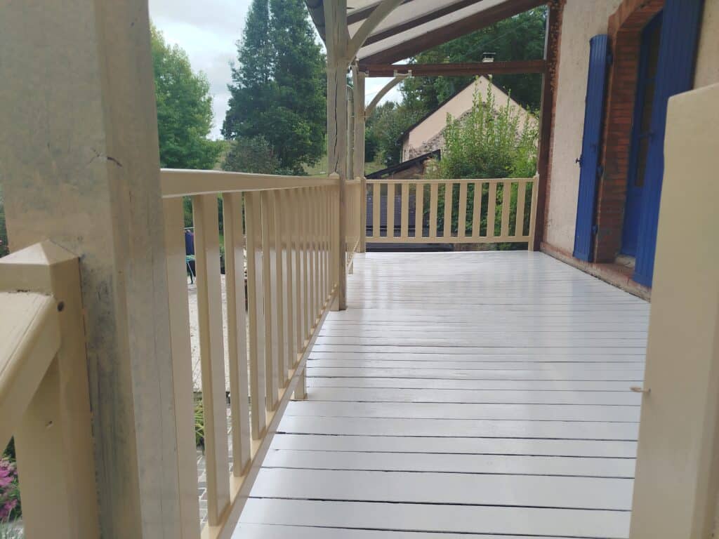 Longue terrasse en bois peinte en blanc avec balustrade beige. Vue sur un jardin verdoyant et une maison aux volets bleus.