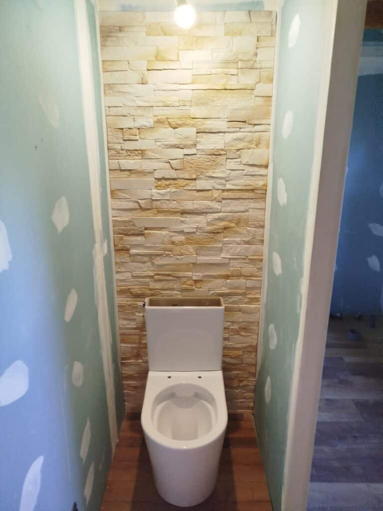 Toilette blanche installée devant un mur texturé en pierre beige dans un petit espace. Les murs latéraux sont en placo en construction.